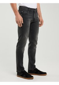 Mężczyzna ubrany w ciemnoszare dżinsy o dopasowanym kroju i czarne trampki z gumowymi podeszwami, stojący na jednolitym jasnym tle.