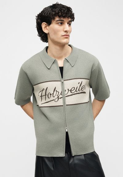 Νέος άνδρας με σγουρά σκούρα μαλλιά, ντυμένος με κοντομάνικο φούτερ ανοιχτού πράσινου χρώματος με φερμουάρ και τη λέξη "Holzweiler" στο στήθος.