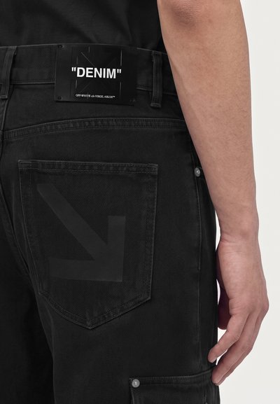 Gros plan d’un pantalon en denim noir avec un patch noir étiqueté "DENIM" et un motif discret de flèche sur la poche arrière, porté par une personne.