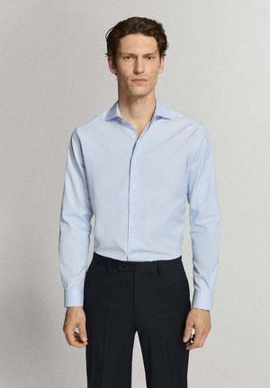 Mann mit dunklen Haaren, der ein hellblaues Hemd mit Knöpfen und schwarze Hose trägt, steht vor einem schlichten hellen Hintergrund.