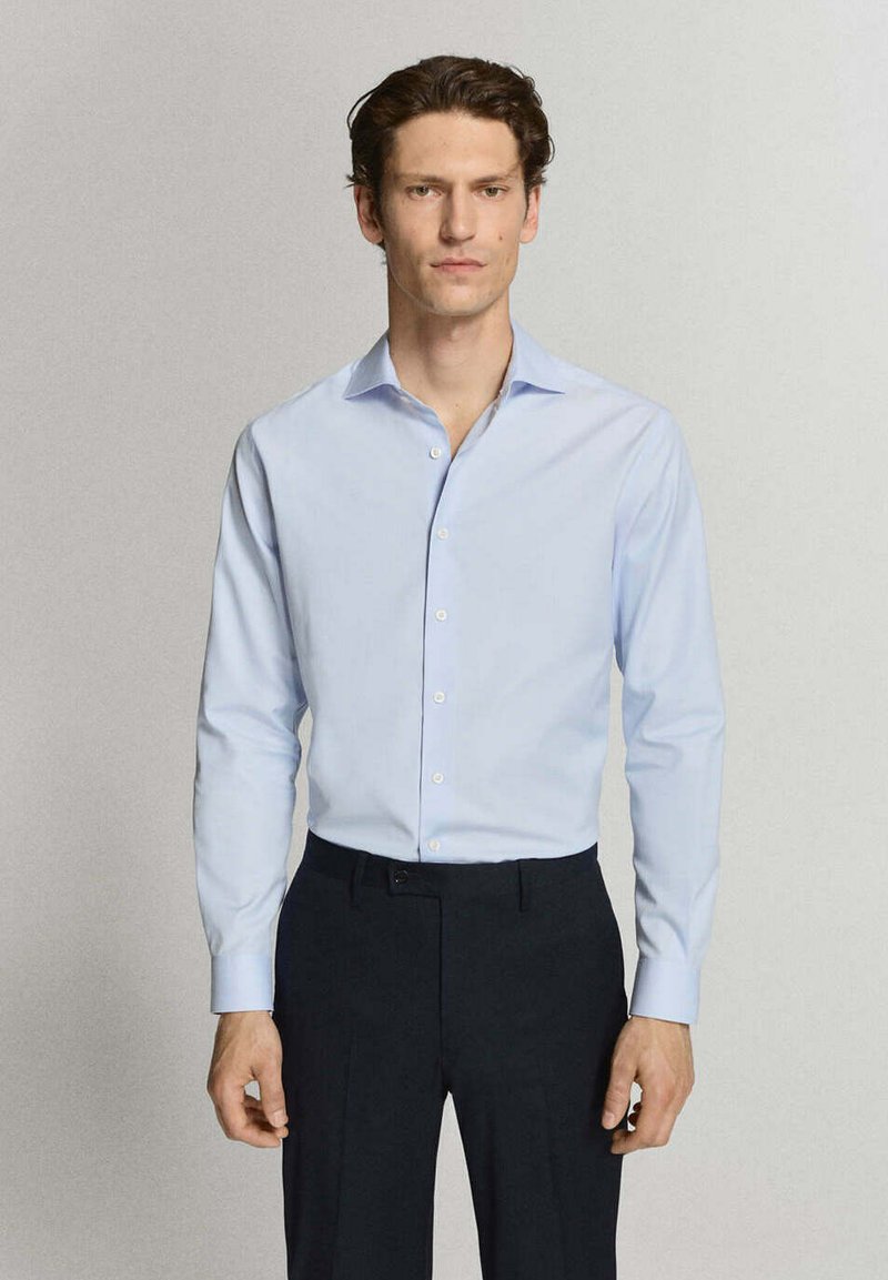 Homme aux cheveux foncés, portant une chemise bleu clair à boutons et un pantalon noir, debout devant un fond clair uni.