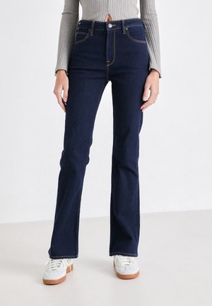 Jeans a zampa d'elefante blu scuro in denim, con vita alta, design a cinque tasche e cuciture in contrasto. Indossati con sneakers bianche.