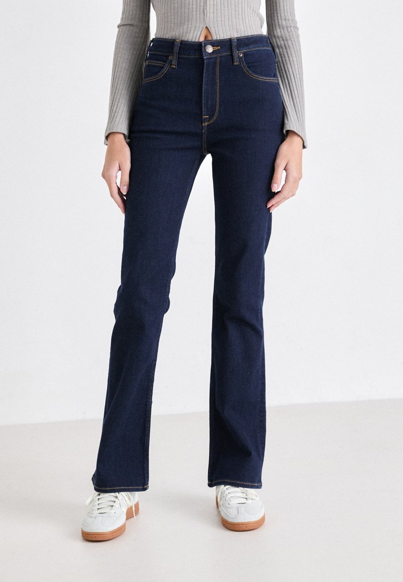 Jeans a zampa d'elefante blu scuro in denim, con vita alta, design a cinque tasche e cuciture in contrasto. Indossati con sneakers bianche.