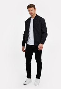 Schwarze Bomberjacke aus glänzendem Material, mit gerippten Bündchen und Kragen, mit Reißverschluss vorne. Getragen über einem weißen T-Shirt, kombiniert mit engen schwarzen Hosen und weißen Sneakers.