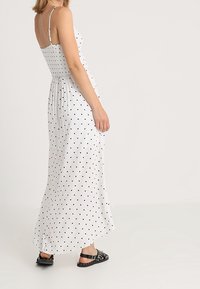 Bílá maxi šaty s černými puntíky, nastavitelné tenké ramínka, elastický pas, splývavá látka a v kombinaci s černými sandály.