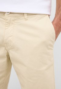 Chinos en coton beige avec une texture lisse, fermeture à bouton et poches latérales, dotés de détails de couture subtils et d'une coupe ajustée.