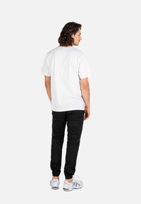 Hombre con cabello de longitud media, vistiendo una camiseta blanca lisa, pantalones jogger negros y zapatillas deportivas blancas, de pie mirando hacia atrás.