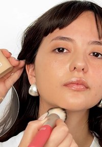 Meikkiä sovelletaan; malli pitää kädessään beigeä meikkipohjaa sisältävää kompaktia, suurta hopeista korvakorua ja vaaleanpunahandleista sivellintä.