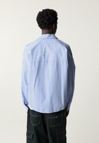 Chemise bleu clair à manches longues avec des rayures verticales blanches et pêche, une coupe décontractée et un détail de couture au dos. Le tissu semble lisse et tissé.