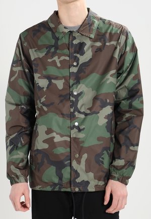 Osoba nosząca zieloną, brązową i czarną kurtkę zapinaną na guziki z elastycznymi ściągaczami i małym logo Nike na piersi.