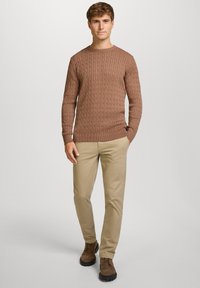 Mężczyzna stojący na tle jednolitego tła, ubrany w brązowy sweter w warkocze, beżowe chinosy i brązowe zamszowe buty z czarną podeszwą.