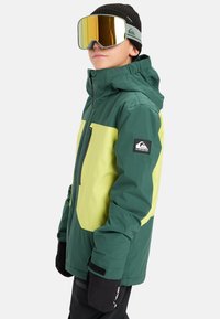 Zielona i żółta zimowa kurtka narciarska z zapięciem na zamek, regulowanymi mankietami i naszywką z logo na rękawie. Model ma na sobie oversize'owe odblaskowe gogle.