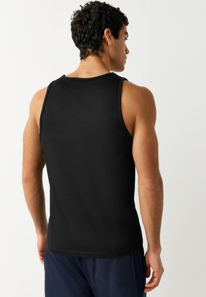 Sötét, göndör hajú férfi, fekete ujjatlan ingben és tengerészkék nadrágban, háttal egy egyszínű világos háttér előtt.