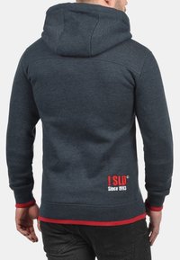 Sudadera gris oscuro hecha de un tejido suave, con capucha, acentos rojos en los puños y el dobladillo, y un parche con el logo en la parte posterior.