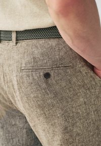 Los pantalones de lino marrones cuentan con un bolsillo trasero plano con un botón negro y un cinturón de acento tejido en verde y blanco. La textura parece suave y ligera.