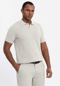 Lys beige kortærmet polo-shirt med en struktureret strik, en blød krave og en to-knaps lukning. Båret med lyse bukser og et sort bælte.