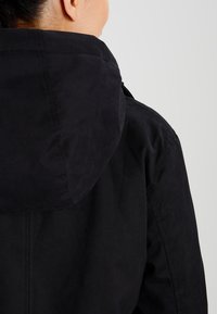 Zwarte jas met capuchon, met een gladde textuur en eenvoudig ontwerp. Naaddetails zichtbaar langs de rug en schouders.