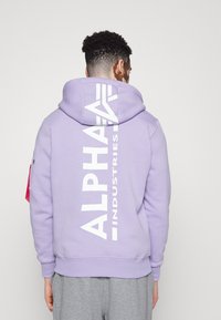 Lavendelfarvet hætte sweatshirt lavet af blødt stof, med et stort hvidt "ALPHA INDUSTRIES" logo trykt vertikalt på bagsiden.