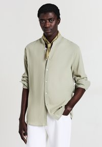 Camicia verde chiaro a maniche lunghe con colletto alla cinese, caratterizzata da un tessuto testurizzato. La camicia ha le maniche arrotolate ed è abbinata a pantaloni bianchi.