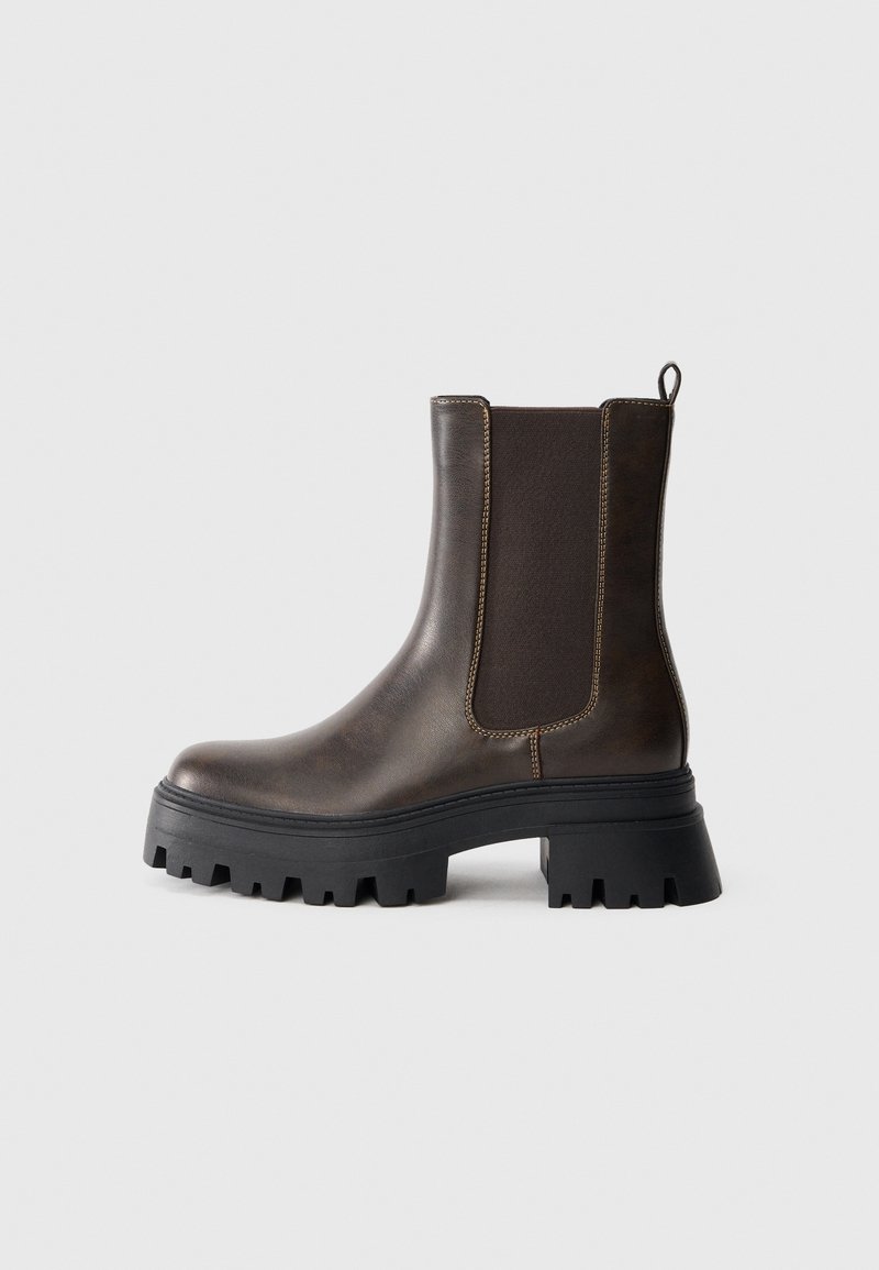 Bottine Chelsea en cuir marron foncé avec semelle épaisse noire à crampons et panneaux latéraux élastiques sur un fond clair uni.