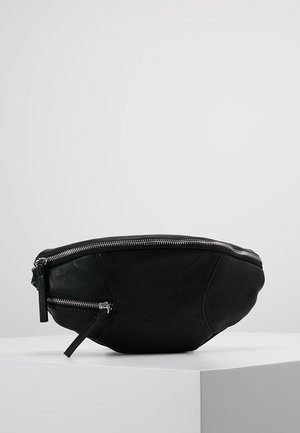 Sac banane en cuir noir avec deux compartiments zippés argentés, posé sur une surface blanche devant un fond gris clair.