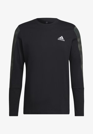 Homme portant un t-shirt Adidas à manches longues noir avec des rayures olive foncé sur les manches et le logo Adidas sur le côté gauche de la poitrine, les mains jointes devant lui.