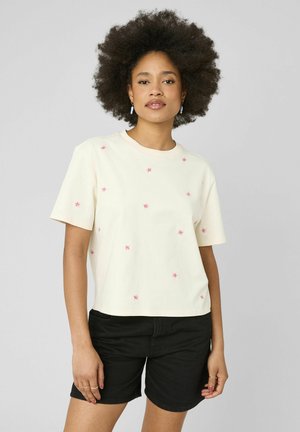 Femme avec coiffure afro portant un t-shirt crème avec de petits motifs floraux roses et un short noir sur un fond gris uni.