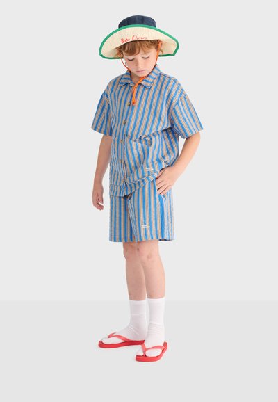 Enfant portant une chemise à manches courtes rayée bleu et orange ainsi qu'un short, chaussettes blanches avec tongs rouges, et un chapeau à large bord regardant vers le bas.
