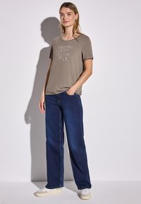 Camiseta de corte holgado en color verde oliva con texto blanco, combinada con jeans de pierna ancha en azul oscuro. El fondo es claro y neutro.