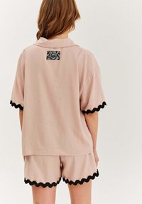 Ružový top s krátkymi rukávmi a čiernym vlnitým lemom a vzorovanou nášivkou na chrbte, spojený s nohavicami so zúbkami v rovnakom štýle.