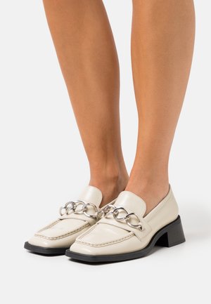 Des mocassins en cuir crème mettent en évidence un accent en chaîne à l'avant et un petit talon carré, avec une finition lisse et des détails de couture.