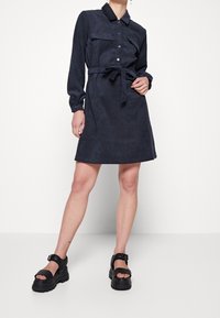 Abito camicia blu navy con maniche lunghe, apertura con bottoni, due tasche sul petto, cintura in tessuto e vestibilità sagomata. Abbinato a sandali neri spessi.