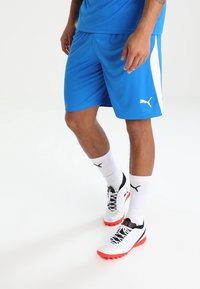 Man som bär blå Puma sportshorts, vita Puma-strumpor och vita och röda Puma sportskor, står på en vit bakgrund.