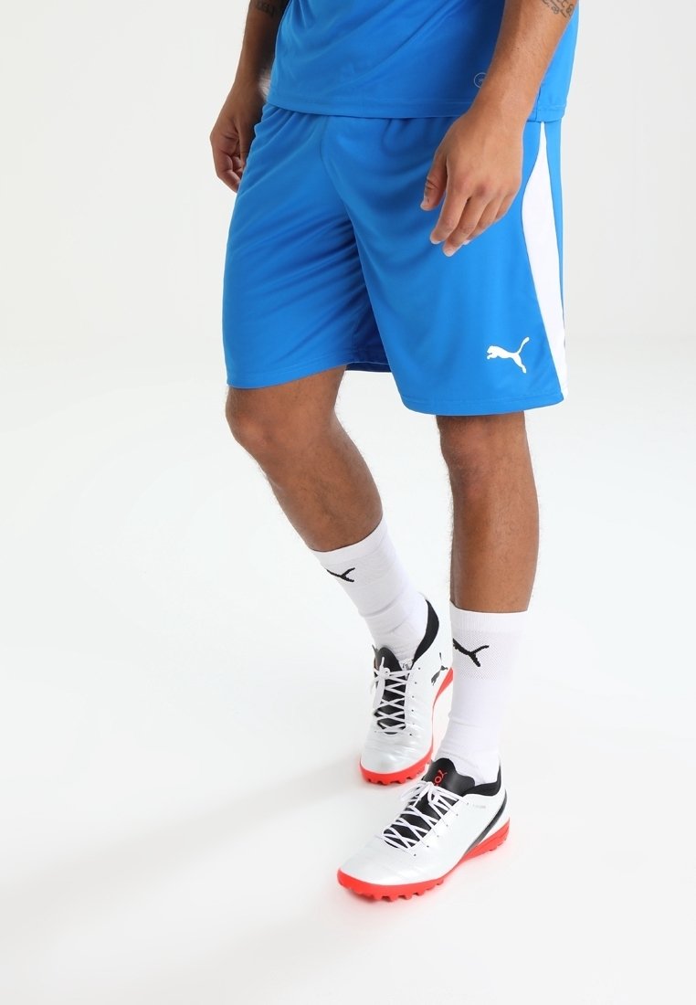 Man som bär blå Puma sportshorts, vita Puma-strumpor och vita och röda Puma sportskor, står på en vit bakgrund.