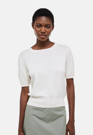 Femme aux cheveux courts portant un haut en maille blanc à manches courtes et une jupe gris clair, debout contre un fond uni clair.