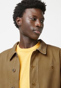 Bruine katoenen jas met knoopsluiting, gelaagd over een gele t-shirt. Heeft een klein logo op de jas en een gestructureerde stof.