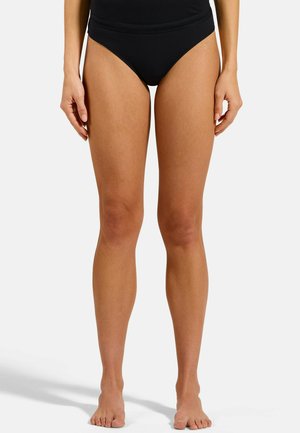 Female legs standing barefoot on a white surface wearing black high-waisted underwear, shown from mid-thigh to feet.