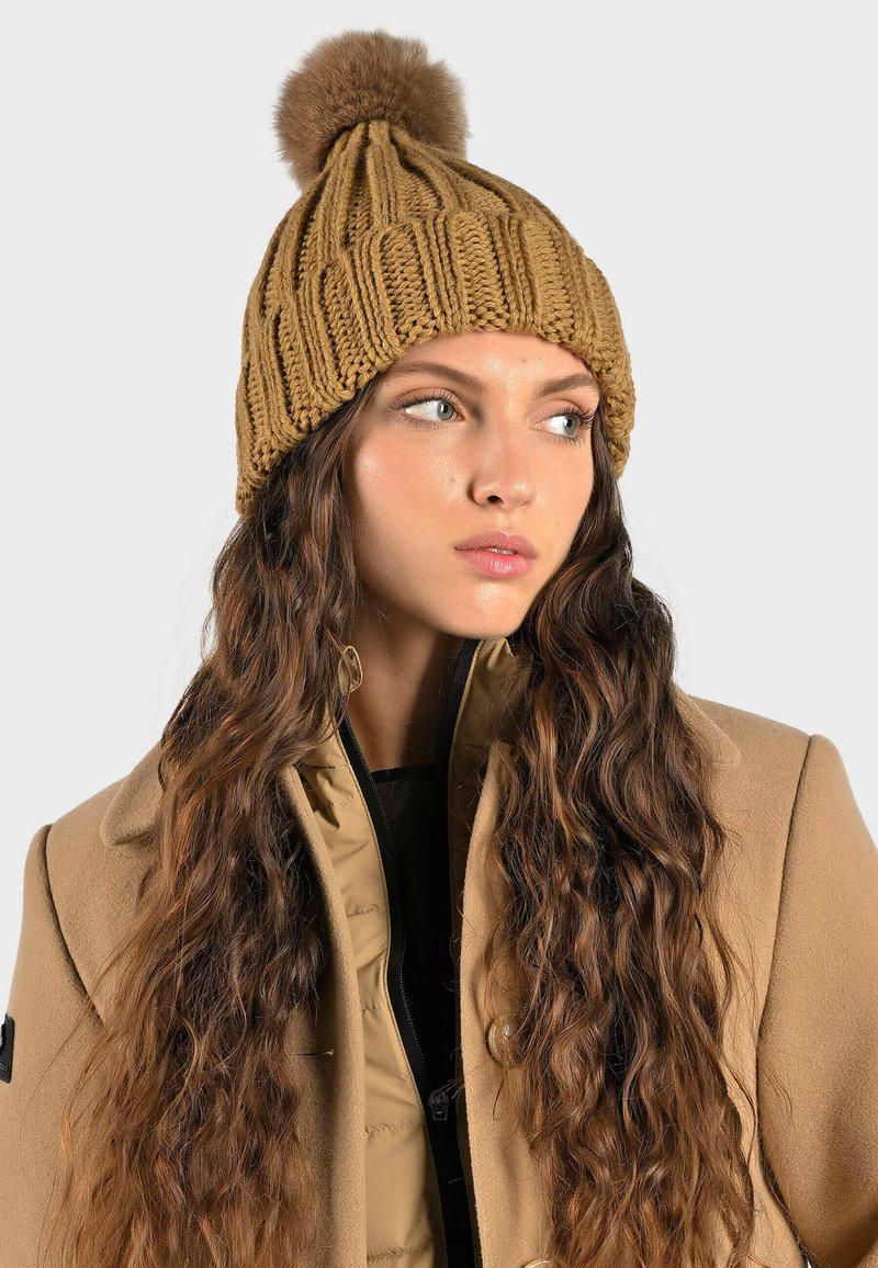 Berretto di lana dorato a maglia con un design a coste testurizzato e un pompon in pelliccia sintetica sulla parte superiore. Indossato con un cappotto beige e presenta capelli lunghi e mossi.
