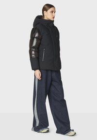 Giacca a vento nera con maniche nere lucide, cappuccio e due tasche frontali con zip, abbinata a pantaloni in denim a gamba larga con strisce laterali grigie.