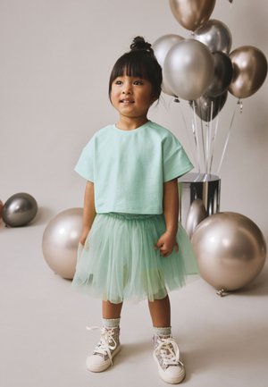 Junges Mädchen trägt ein mintgrünes Oberteil und einen Tüllrock, steht neben metallischen Luftballons in Silber- und Bronzetönen vor neutralem Hintergrund.