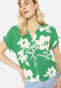 Blusa verde a maniche corte con grandi stampe floreali bianche, abbottonatura frontale e vestibilità morbida. Tessuto leggero e liscio.