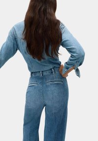 Jumpsuit in denim azzurro chiaro con un corpo aderente, maniche lunghe e pantaloni a gamba larga. Dispone di due tasche posteriori e polsini delle maniche con bottoni.