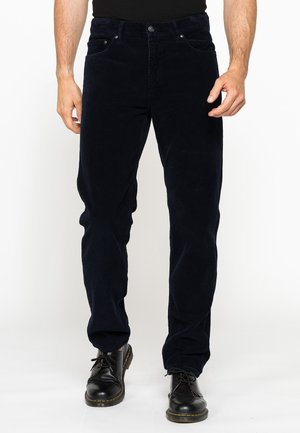 Uomo con pantaloni di velluto a coste neri e scarpe nere stringate, in piedi davanti a uno sfondo bianco uniforme.