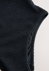 Fekete texturált fürdőruha mintás, gyűrött felülettel. Sima belső bélés és kerek nyakkivágás. Közeli kép az anyagról és a kidolgozásról.