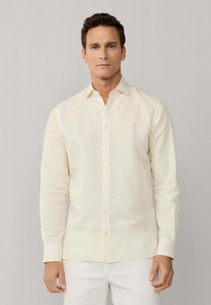 Mann mit kurzen dunklen Haaren trägt ein hellcremefarbenes, langärmliges Hemd mit Knöpfen und weiße Hosen vor einem schlichten beigen Hintergrund.