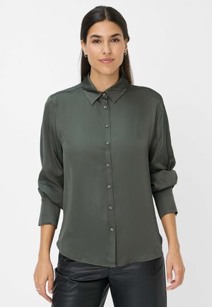 Blusa verde scuro a maniche lunghe con bottoni frontali, colletto e una texture morbida e liscia. Presenta maniche arrotolate e bottoni abbinati al tessuto.