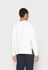 Sudadera blanca con un ajuste relajado, hombros caídos y aberturas laterales. Textura suave, diseño de cuello redondo, combinada con pantalones azul marino oscuros.