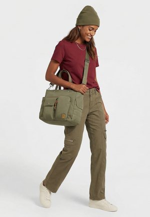 Kvinde iført en rød T-shirt, olivengrønne bukser, hue og hvide sneakers, der bærer en olivengrøn skuldertaske med flere lommer.