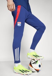Blå idrottsbyxor med röda sidopaneler, med Olympique Lyonnais-logotyp. I kombination med klara gula fotbollsdojor på en färgglad fotboll.