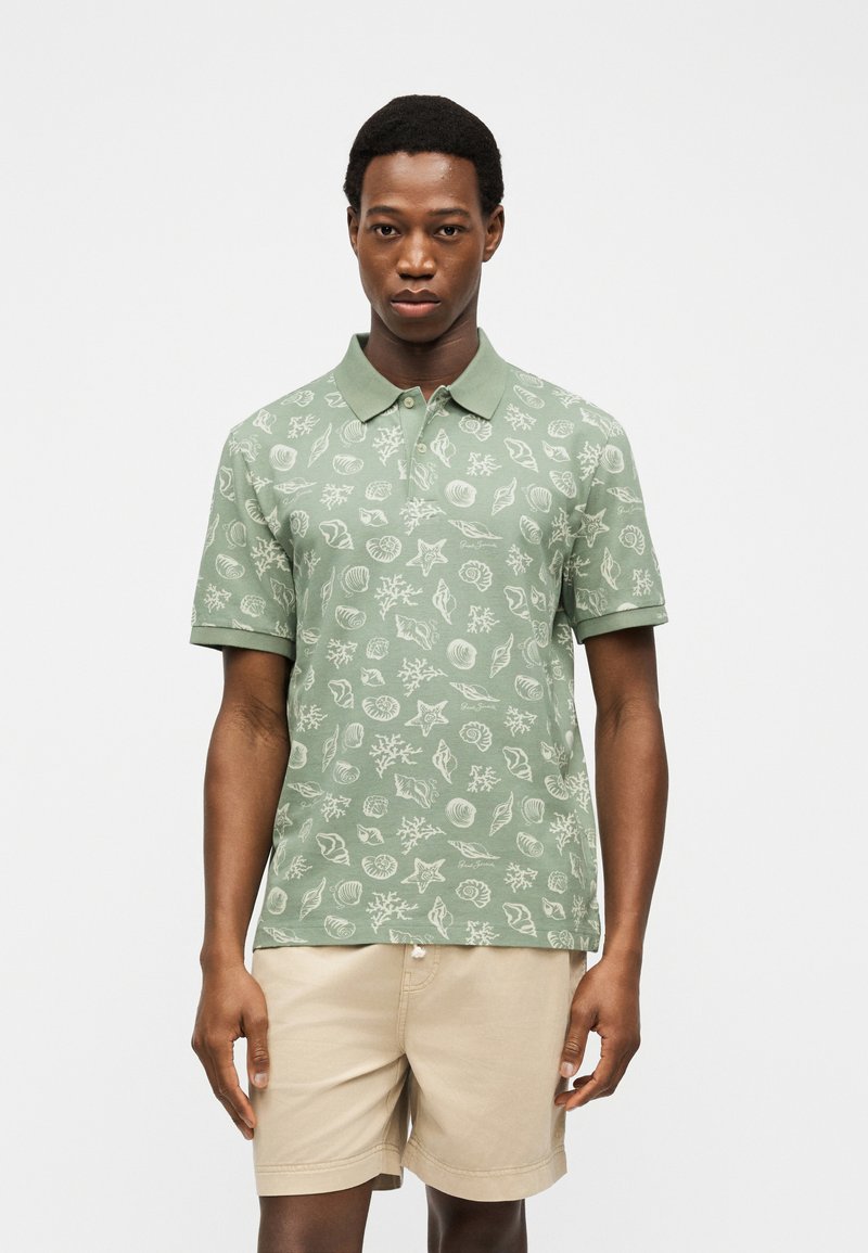 Uomo che indossa una polo verde salvia con motivi di conchiglie bianche e coralli, abbinata a pantaloncini beige, in piedi davanti a uno sfondo semplice.